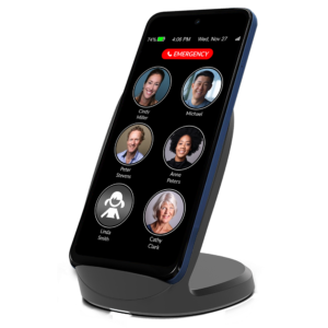RAZ Memory Cell Phone resting upright on a sleek wireless charging stand, showing a simple home screen with large photo buttons for contacts and a prominent red ‘Emergency’ button at the top.