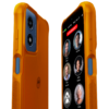Angled view of the RAZ Memory Cell Phone inside the orange Memory Cell Phone Rugged Protective Case. The front screen shows large contact buttons with names and photos—Cindy Miller, Michael, Peter Stevens, Anne Peters, Linda, and Cathy—along with a red “EMERGENCY” button at the top. The back and side of the case are visible, featuring a textured surface, grip-enhancing side panels, and easy-to-press side buttons.